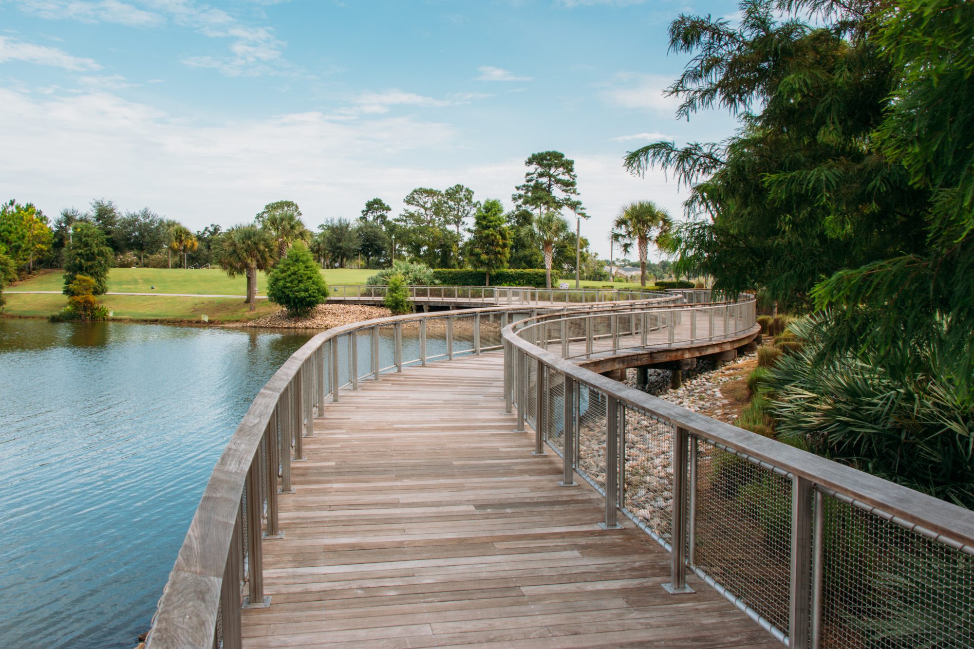 A wooden boardwalk with metal railings curves over a lake, surrounded by trees and greenery, under a partly cloudy sky.