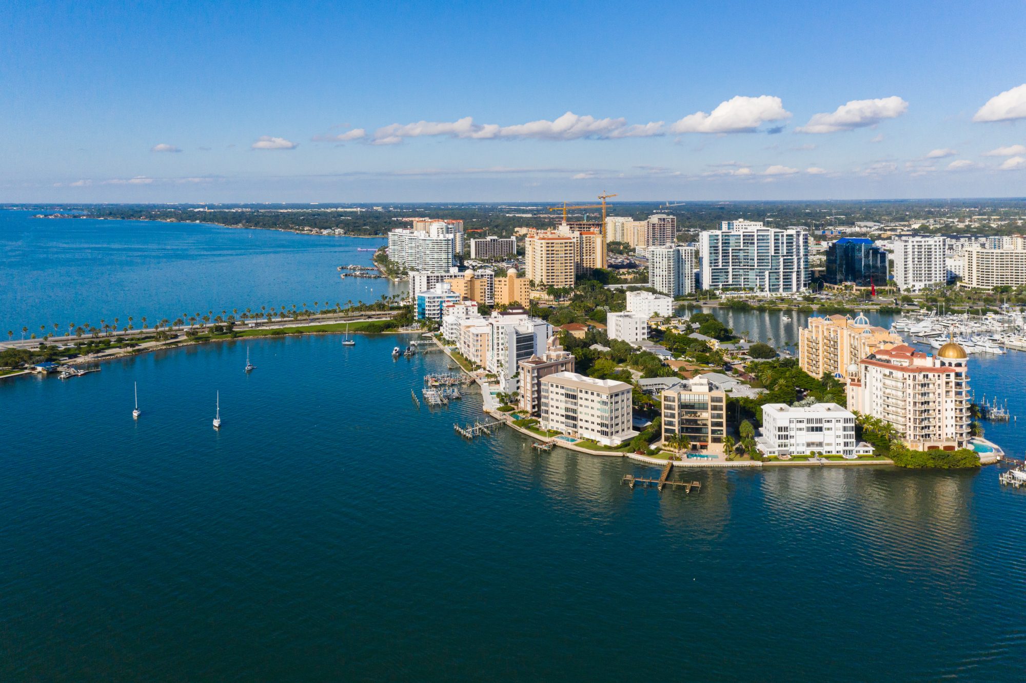 Aerial view of a coastal city with numerous buildings along the shore, a marina with several boats docked, and a body of water extending into the distance under a clear blue sky, showcasing an idyllic scene reminiscent of Reno Living Trust locales.