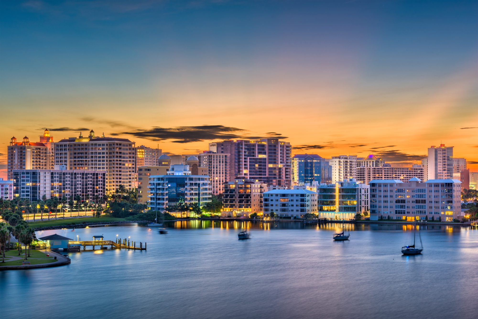 City skyline at sunset with several high-rise buildings, a calm body of water, and small boats anchored near the shore; a serene backdrop for reflecting on your Reno Living Trust or Carson City Estate Planning.