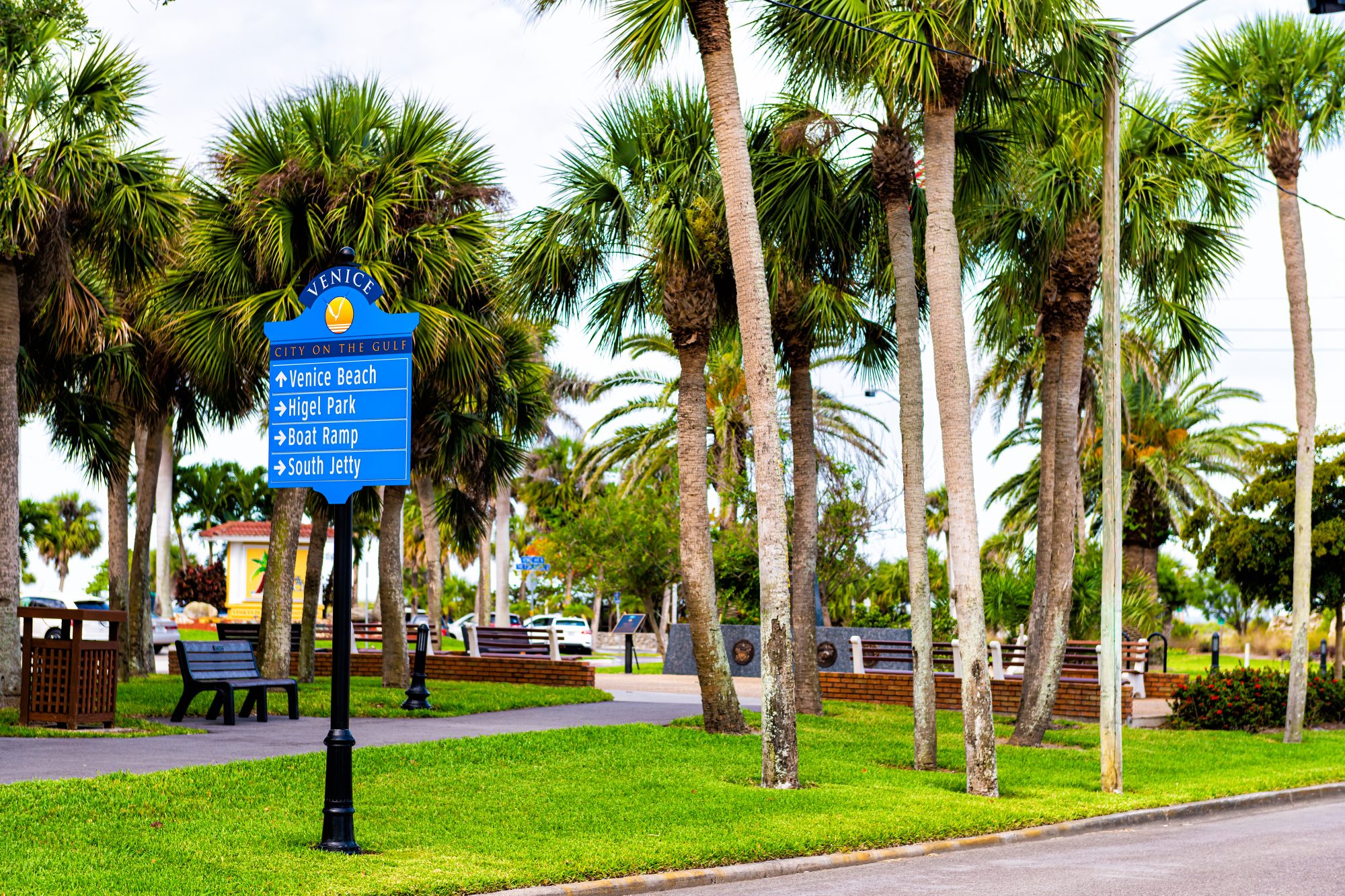 Signpost surrounded by palm trees directing to Venice Beach, Angler Park, Boat Ramp, and South Jetty in a park setting. Benches and a paved path are visible, offering a serene environment perfect for contemplating important matters like Carson City Estate Planning.