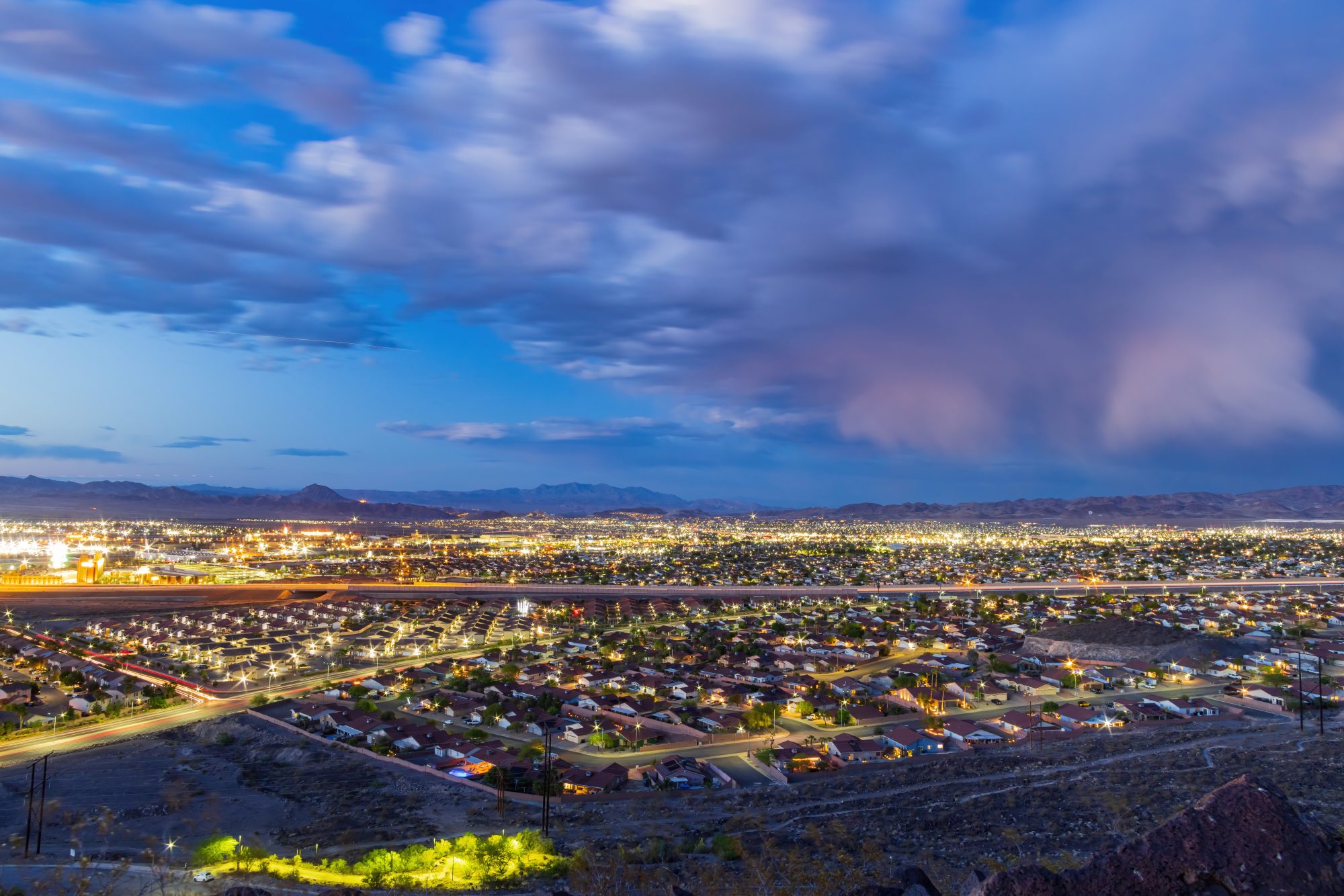 A vast cityscape with glowing lights under a cloudy dusk sky, showcasing a blend of urban buildings, streets, and a distant mountain range; a perfect backdrop for those considering Reno Living Trust options.