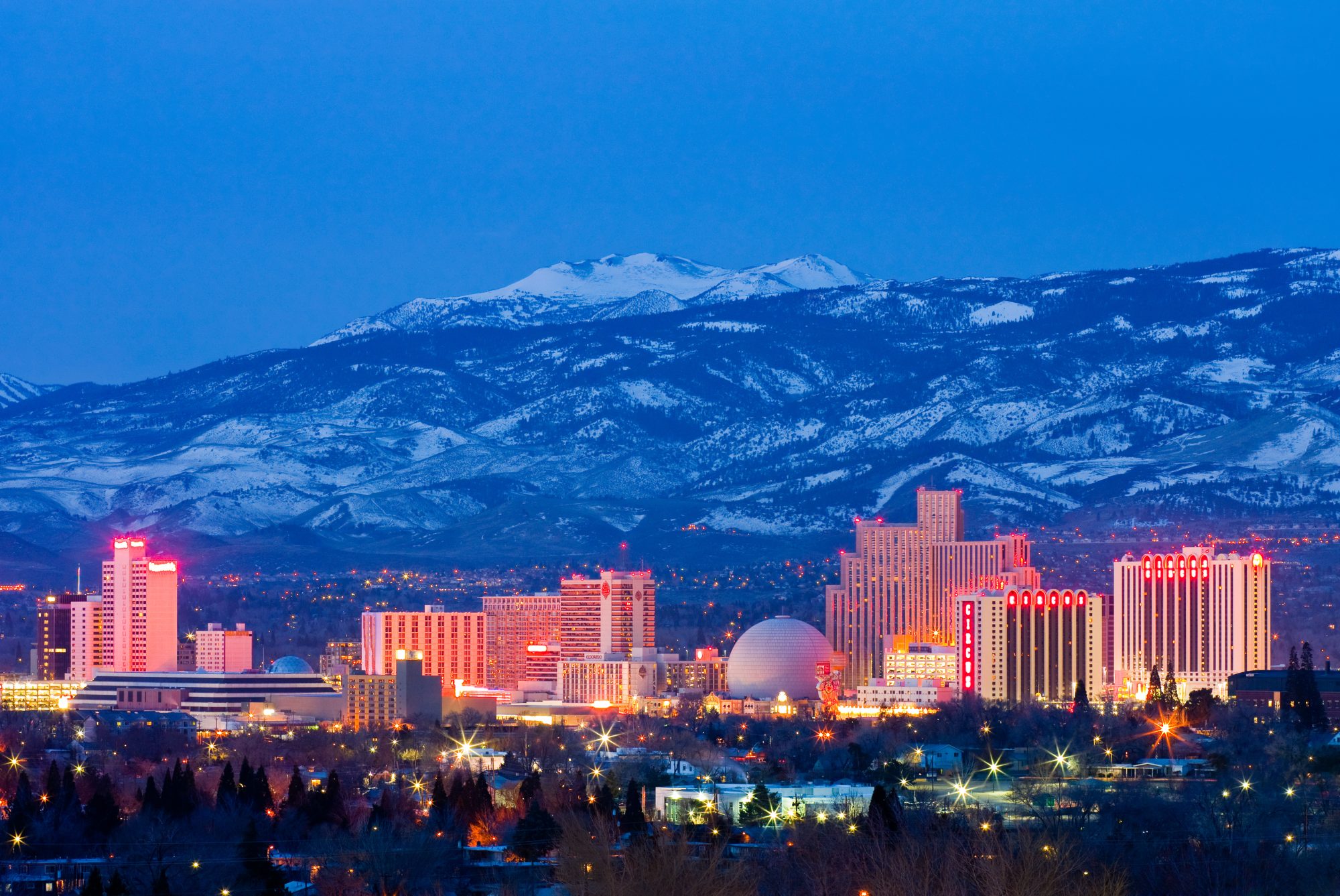 A nighttime view of Reno, Nevada with illuminated city buildings in the foreground and snow-capped mountains in the background, showcasing an area where Carson City Estate Planning is key for residents admiring both urban and natural beauty.