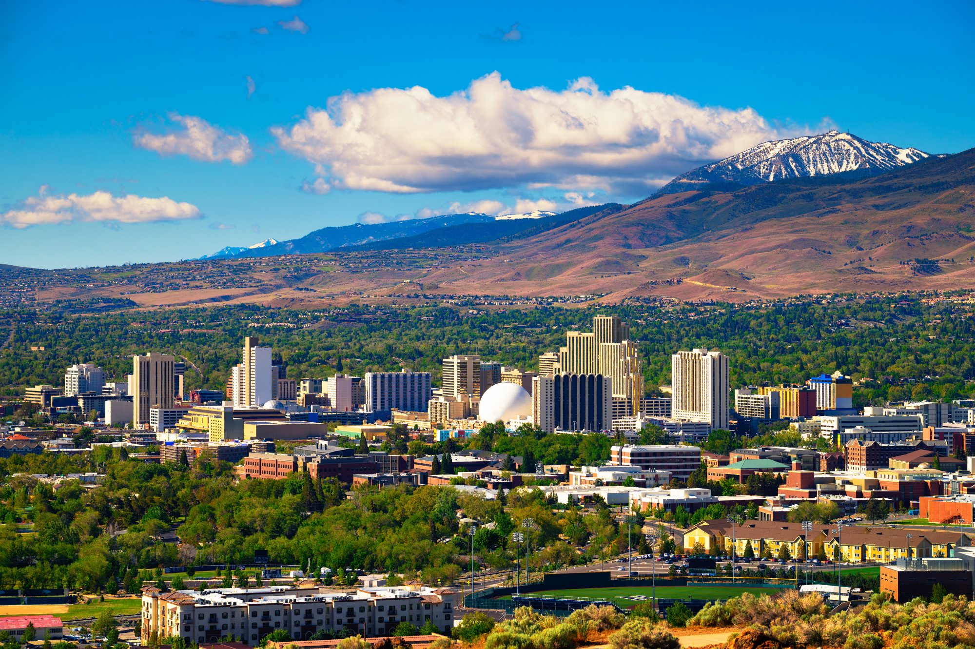 A city skyline featuring tall buildings is set against a backdrop of mountains, with a mix of green foliage and urban structures under a blue sky with scattered clouds, perfectly embodying the blend of natural beauty and modern convenience that Verdi Affordable Living Trust offers.