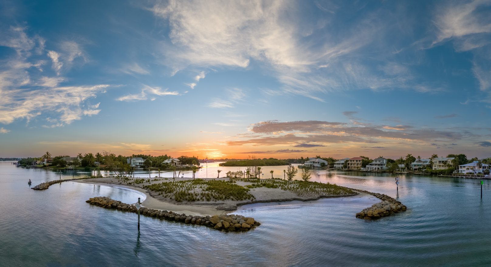 Aerial view of a small island with vegetation surrounded by calm water at sunset. Several houses, perhaps part of properties managed under the Sparks Revocable Living Trust, are visible along the shoreline in the background.