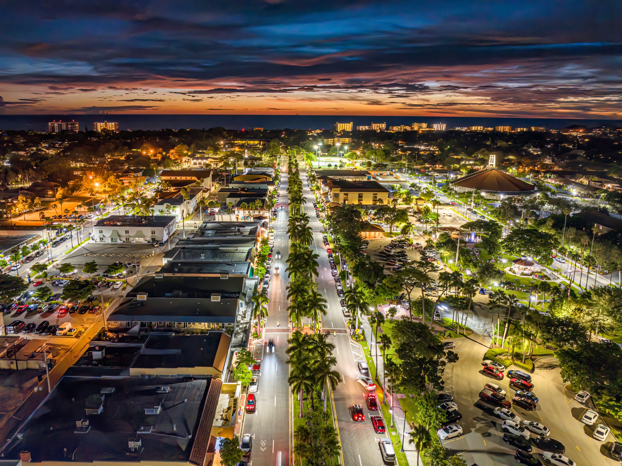 Aerial view of a brightly lit city street at night with buildings, cars, and trees lining the road, and a colorful sunset in the background, showcasing the vibrant life near Reno Living Trust offices.