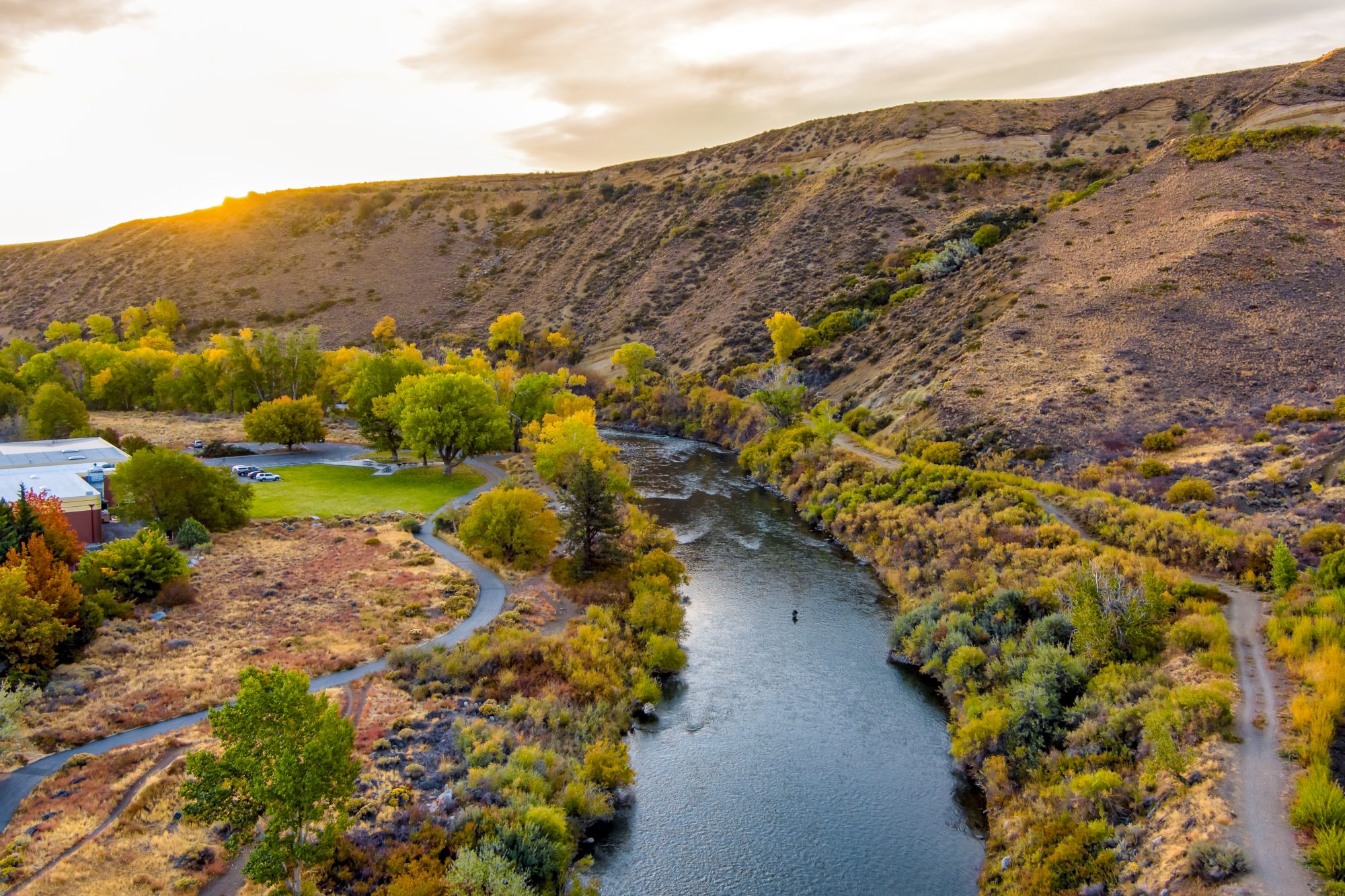 A serene river flows through a hilly landscape with trees in fall colors and a grassy area, where a few parked cars suggest Verdi Affordable Living Trust properties. The sun sets behind the hills, casting a warm glow.