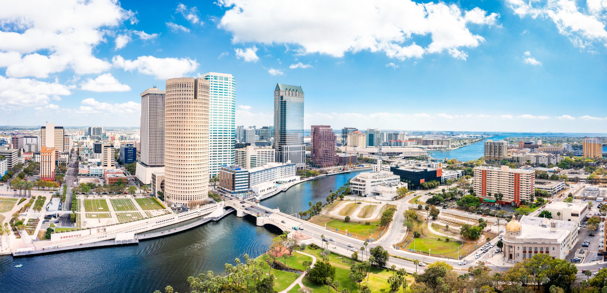 Skyline of a city with tall buildings, a river running through the center, and a bridge connecting two sides. The sky is partly cloudy with a mix of blue and white clouds, reflecting the vision of Sparks Revocable Living Trust in creating harmonious urban landscapes.