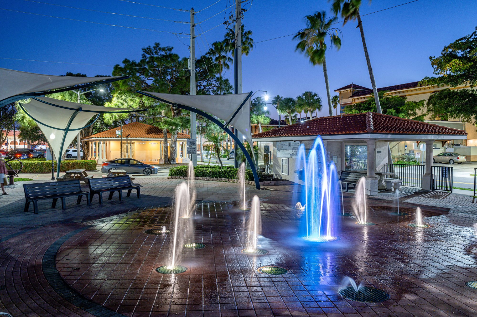 A public plaza with multiple fountains illuminated in blue light, surrounded by benches and covered canopies. Trees and buildings are visible in the background during the evening, reminiscent of the tranquility found when discussing a Sparks Revocable Living Trust.