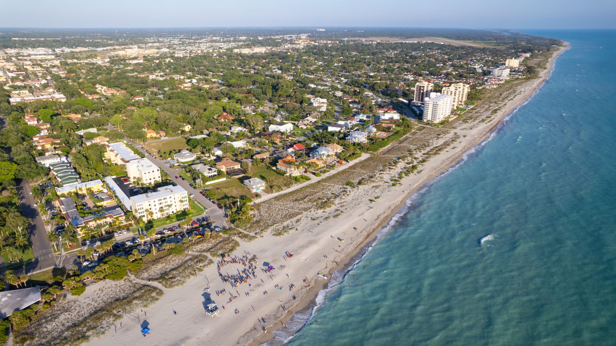 Aerial view of a coastal town with residential buildings and a populated beach, bordered by greenery on one side and the ocean on the other.