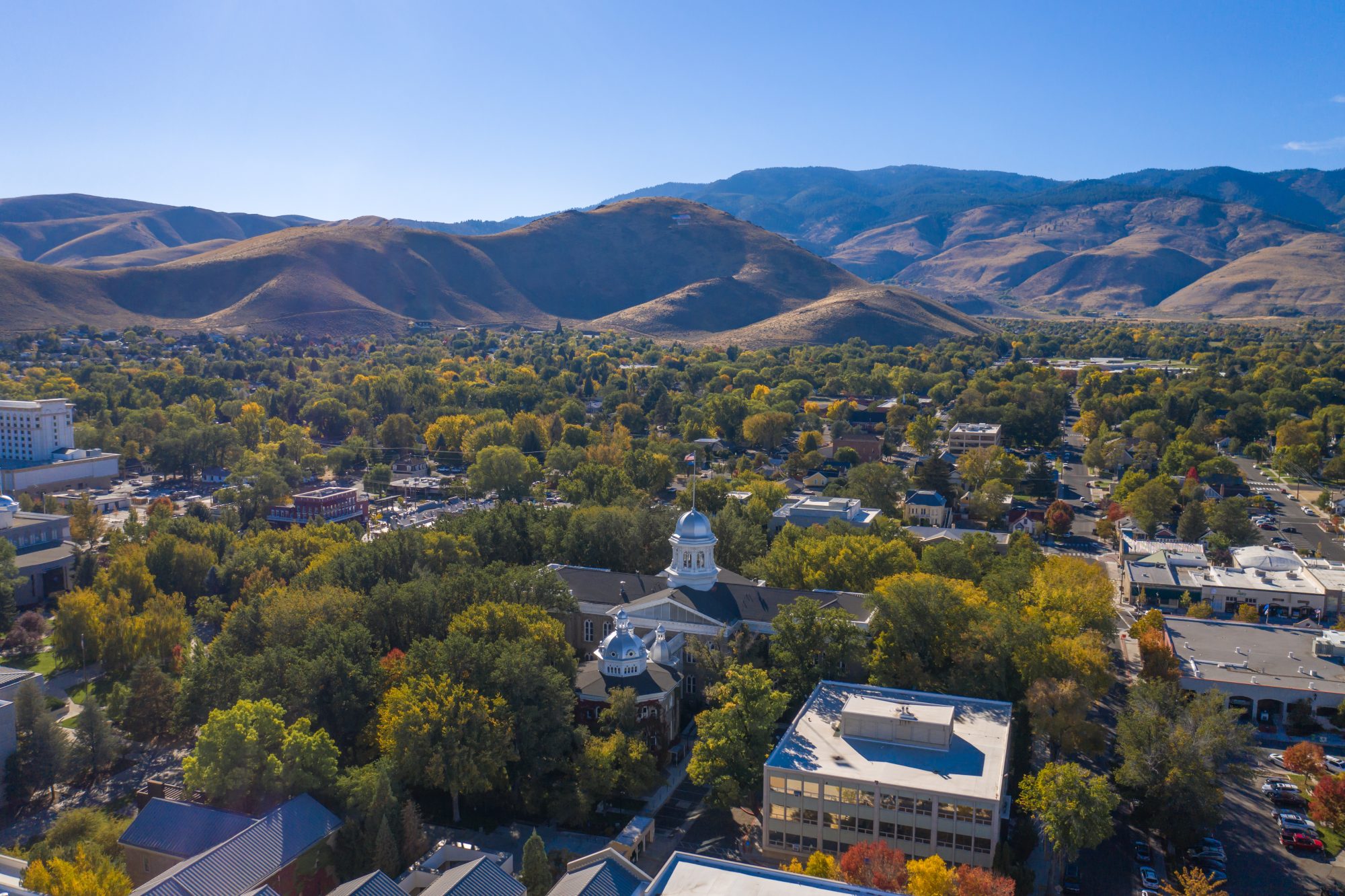 Aerial view of a town surrounded by hills, featuring a white-domed building among trees. Clear blue sky in the background.